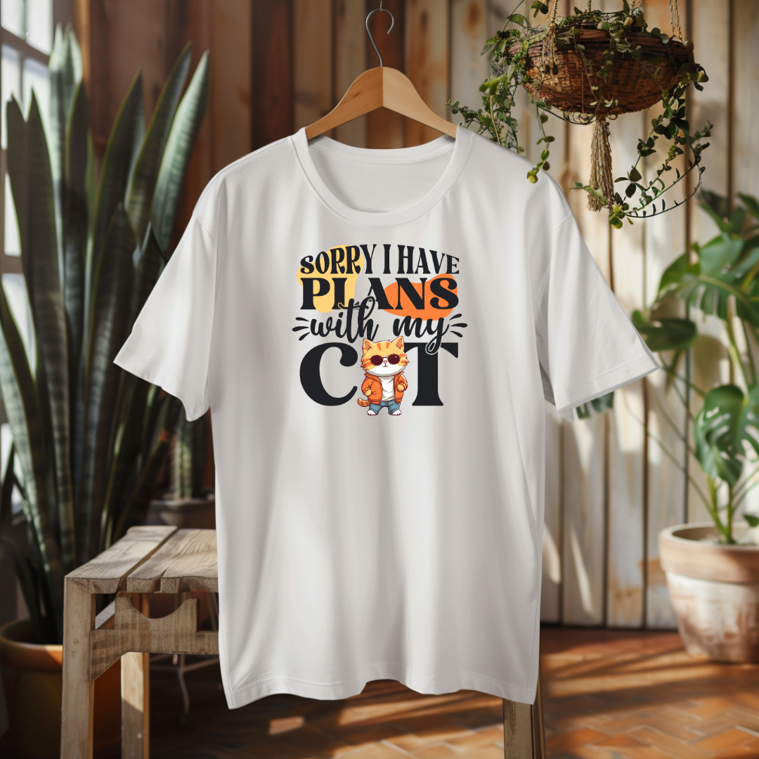 A white t-shirt with a cat-themed design hanging on a wooden hanger in a room with plants.