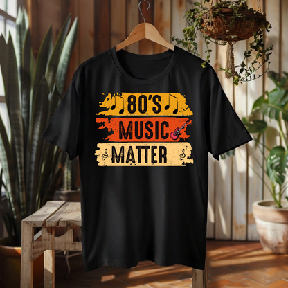 A black short-sleeve t-shirt hanging on a wooden hanger against a rustic wood-paneled wall with plants and a bench. The shirt features a distressed graphic that reads '80's MUSIC MATTER' in black text over yellow, orange, and beige horizontal stripes.