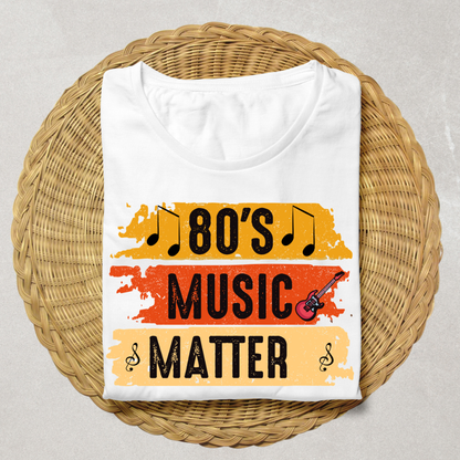A folded white t-shirt with a retro graphic reading '80's MUSIC MATTER' in black text over orange and beige distressed paint strokes, displayed on a round woven wicker mat against a light gray background.