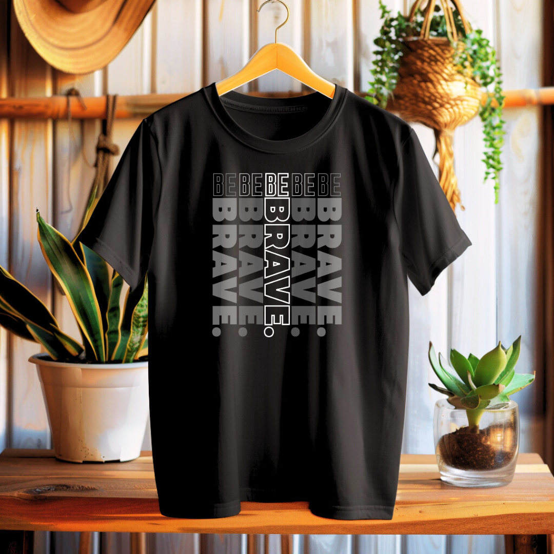 Black t-shirt with 'Be Brave' text on a wooden surface with plants in the background