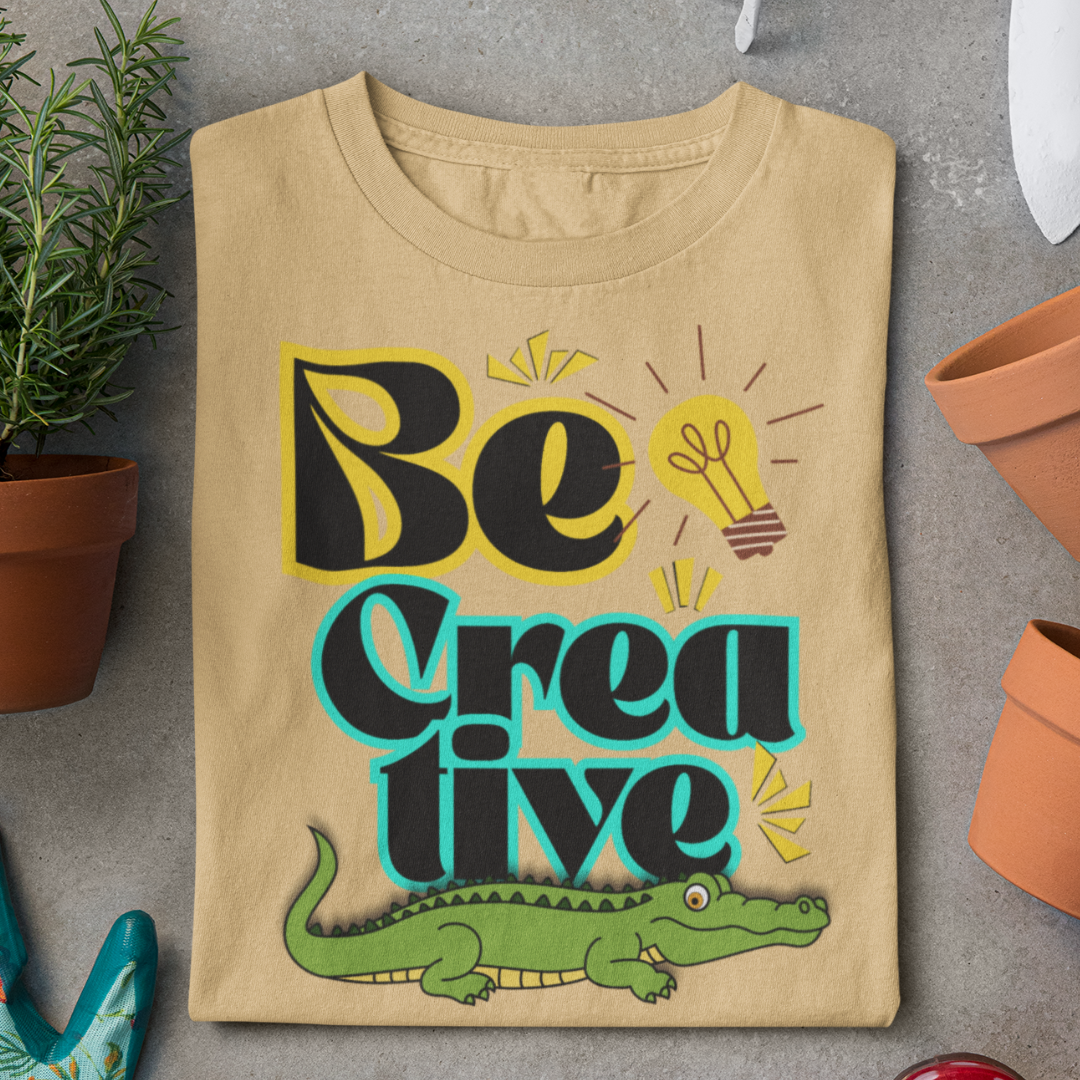 Beige t-shirt with 'Be Creative' text and alligator graphic on a gray surface.