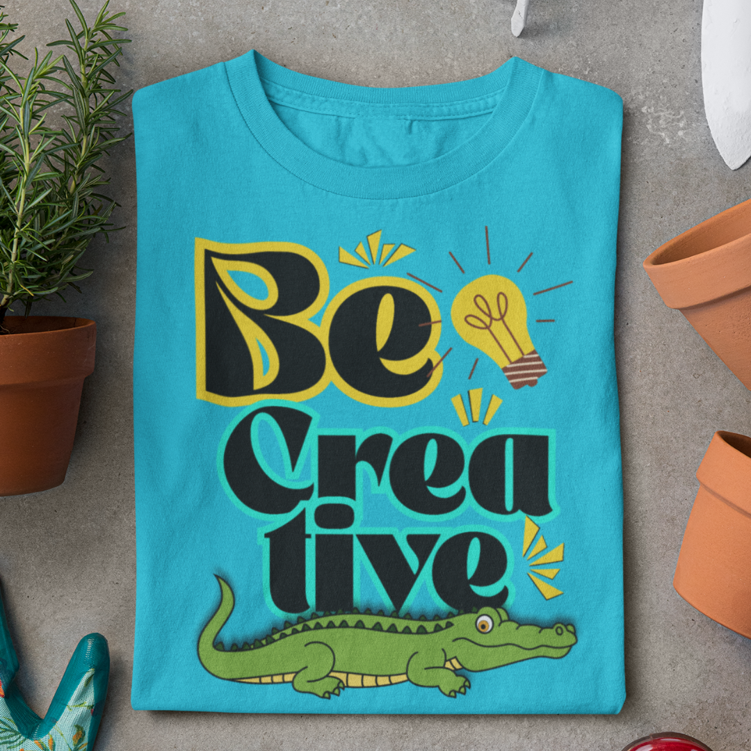 Blue t-shirt with 'Be Creative' design featuring a crocodile, on a gray surface with plants.