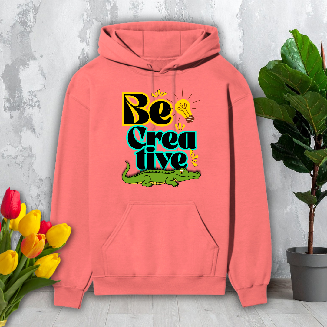 Pink hoodie with 'Be Creative' text and alligator graphic on a light gray surface with flowers and a plant.