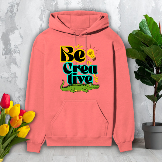 Pink hoodie with 'Be Creative' text and alligator graphic on a light gray surface with flowers and a plant.