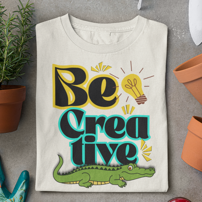 White t-shirt with 'Be Creative' design featuring a crocodile, on a gray surface with plants.
