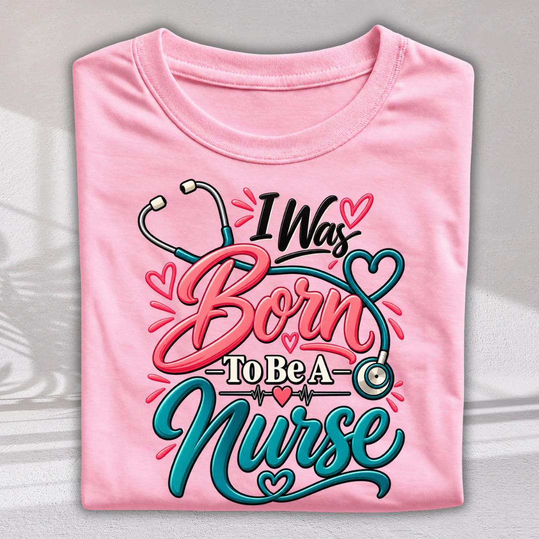 A folded light pink t-shirt with a colorful graphic that reads 'I Was Born To Be A Nurse' in stylized pink and teal lettering, decorated with stethoscope and heart motifs, shown against a neutral gray background.