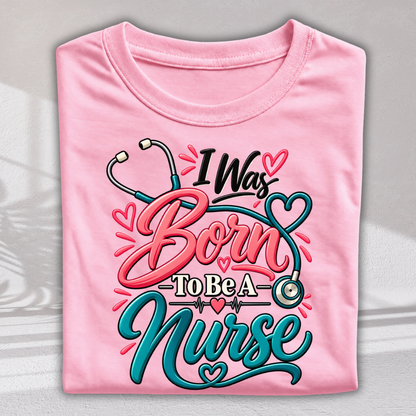 A folded light pink t-shirt with a colorful graphic that reads 'I Was Born To Be A Nurse' in stylized pink and teal lettering, decorated with stethoscope and heart motifs, shown against a neutral gray background.