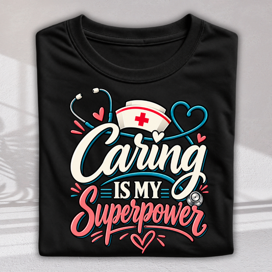 A black folded T-shirt with a colorful graphic that reads 'Caring is my superpower' in white, pink, and blue lettering, decorated with a nurse hat, stethoscope, and hearts, shown on a light gray background.