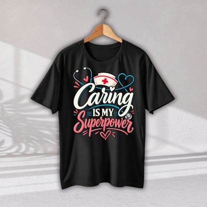 A black short-sleeve t-shirt hanging on a wooden hanger against a light gray wall. The shirt features a graphic print on the front with the text 'Caring is my Superpower' in white and pink lettering, decorated with a nurse hat, stethoscope, and heart motifs.