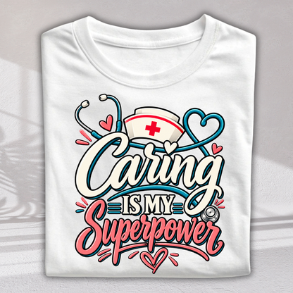 A folded white t-shirt with a colorful graphic print that reads 'Caring IS MY Superpower' in stylized lettering, decorated with a stethoscope, nurse hat, and hearts.