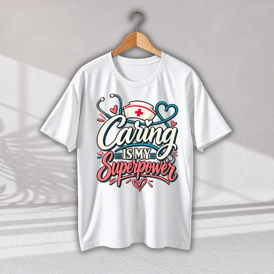 A white short-sleeve crewneck t-shirt hanging on a wooden hanger against a light gray wall. The shirt features a colorful graphic on the front with the text 'Caring IS MY Superpower' in stylized white, pink, and blue lettering, decorated with a nurse hat, stethoscope, and hearts.