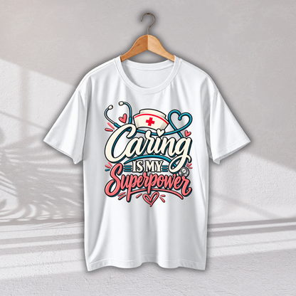 A white short-sleeve crewneck t-shirt hanging on a wooden hanger against a light gray wall. The shirt features a colorful graphic on the front with the text 'Caring IS MY Superpower' in stylized white, pink, and blue lettering, decorated with a nurse hat, stethoscope, and hearts.