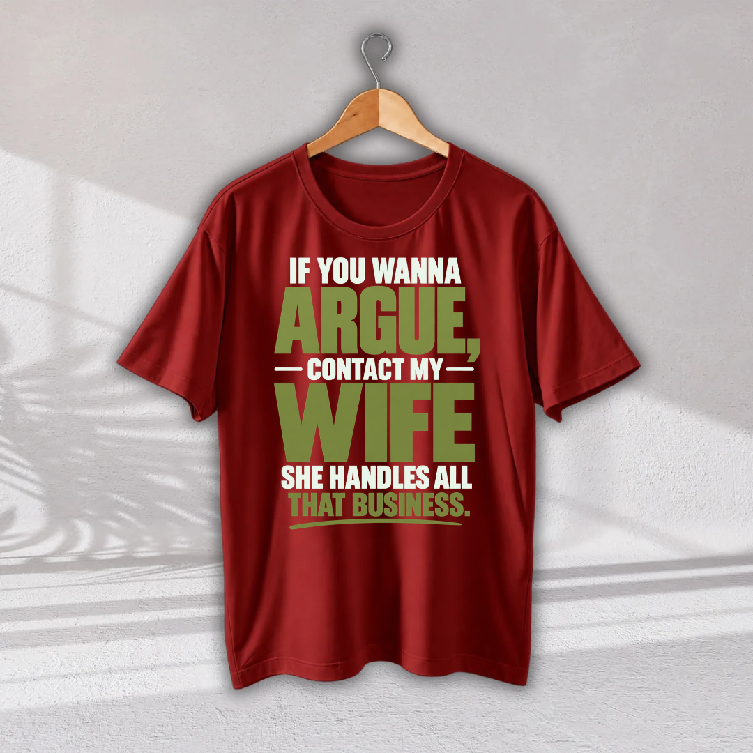A red short-sleeve graphic T-shirt hanging on a wooden hanger against a light gray wall. The shirt features a bold text graphic in white and olive green that reads, 'IF YOU WANNA ARGUE, CONTACT MY WIFE SHE HANDLES ALL THAT BUSINESS.