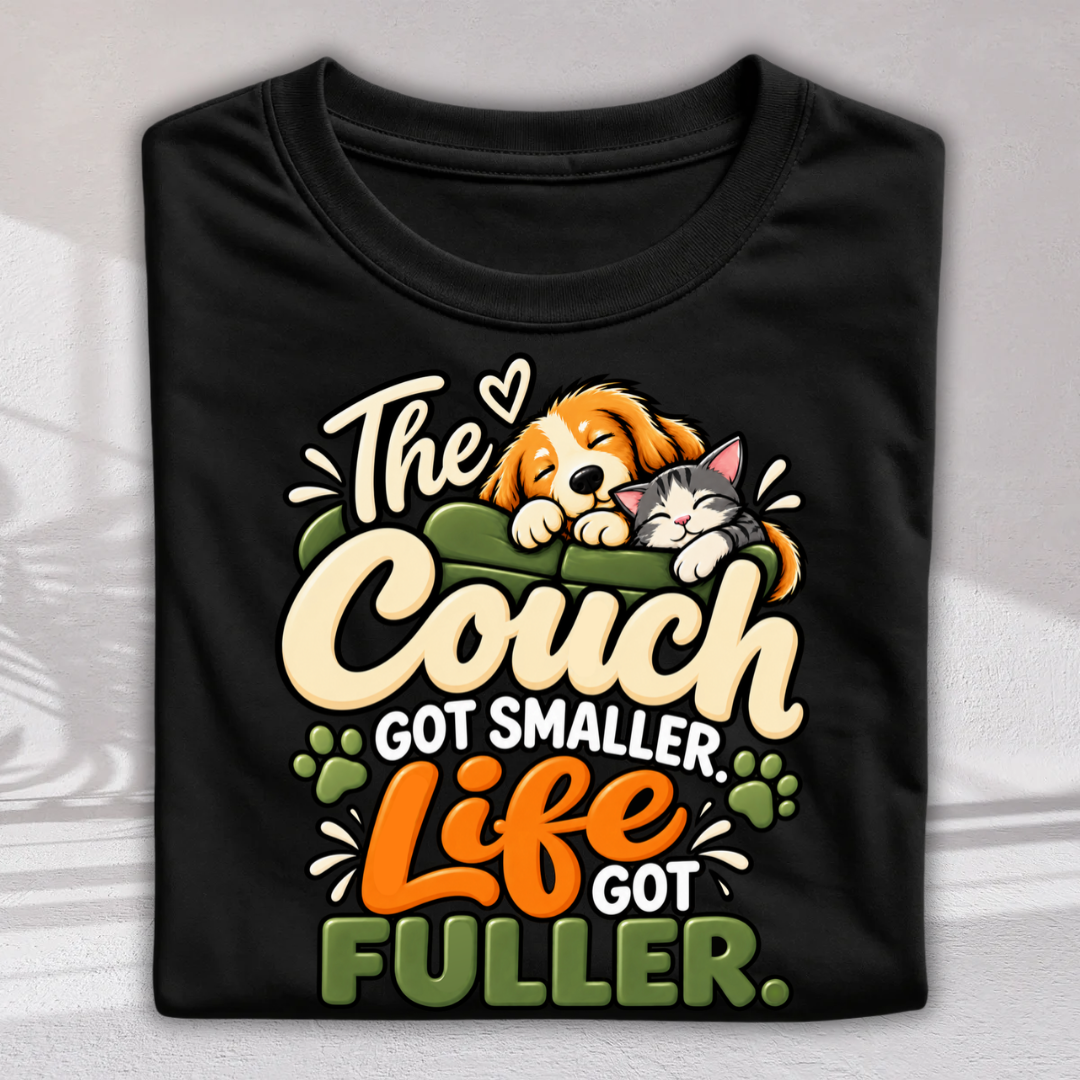 A folded black t-shirt with a colorful graphic print featuring a cartoon dog and cat on a couch and the text 'The Couch GOT SMALLER, Life GOT FULLER.'.