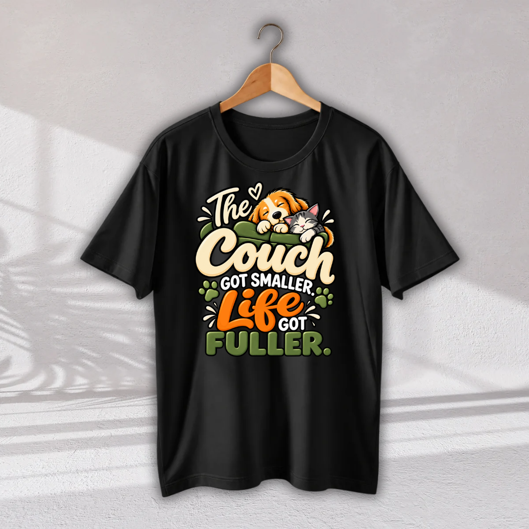 A black short-sleeve graphic T-shirt hanging on a wooden hanger against a light gray wall. The shirt features a colorful front print with the phrase 'The Couch GOT SMALLER Life GOT FULLER.