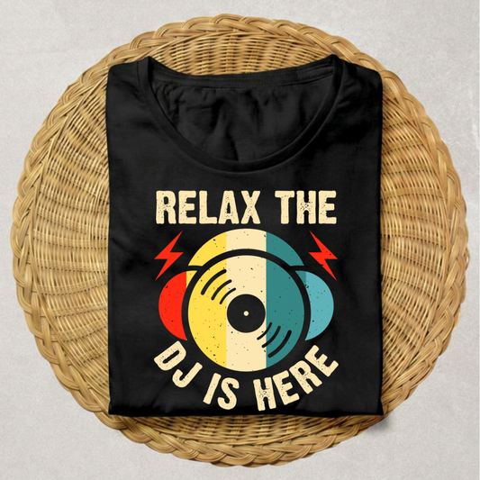A black t-shirt with a retro graphic design featuring a vinyl record and the text 'RELAX THE DJ IS HERE' in cream, red, and teal, displayed flat on a round woven wicker tray.