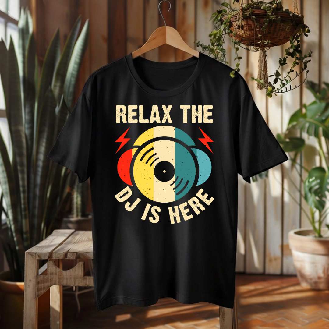 A black short-sleeve t-shirt hanging on a wooden hanger in a rustic indoor setting with plants and wood paneling. The shirt features a retro-style graphic of a vinyl record with red, yellow, and blue sections, flanked by red lightning bolts, with the text 'RELAX THE DJ IS HERE' in cream lettering.