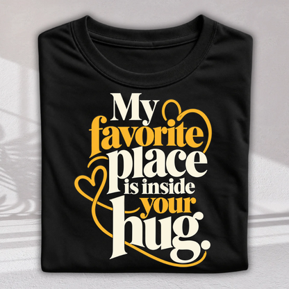 A folded black t-shirt with a graphic print that reads 'My favorite place is inside your hug.' in white and yellow lettering with heart outlines, displayed on a light gray background.