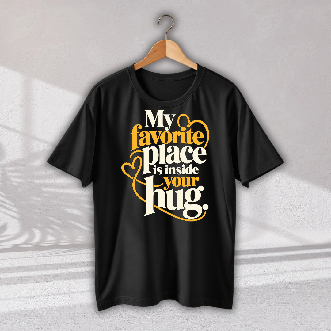 A black short-sleeve t-shirt hanging on a wooden hanger against a light gray wall. The shirt features a graphic print on the front with the text 'My favorite place is inside your hug.