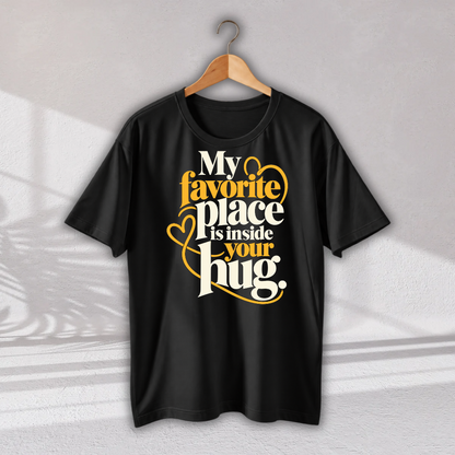 A black short-sleeve t-shirt hanging on a wooden hanger against a light gray wall. The shirt features a graphic print on the front with the text 'My favorite place is inside your hug.