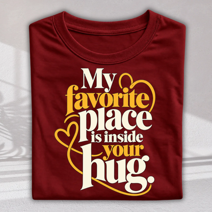 A folded maroon crewneck t-shirt with a graphic print that reads 'My favorite place is inside your hug.' in white and yellow lettering with heart and swirl accents, displayed on a light gray background.