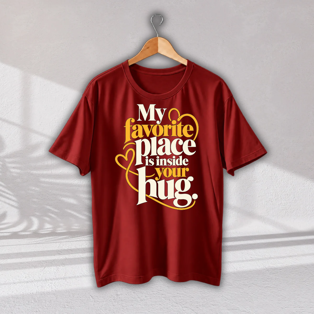 A red short-sleeve t-shirt hanging on a wooden hanger against a light gray wall. The shirt features a graphic print with the text 'My favorite place is inside your hug.