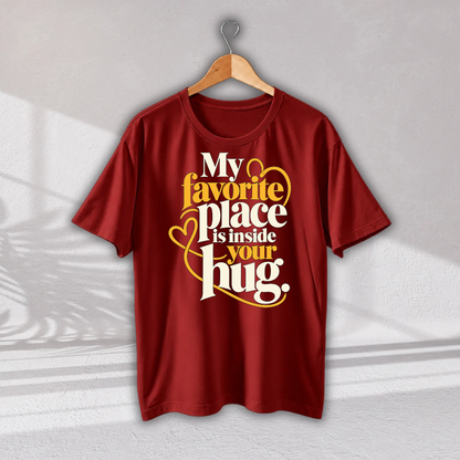 A red short-sleeve t-shirt hanging on a wooden hanger against a light gray wall. The shirt features a graphic print with the text 'My favorite place is inside your hug.