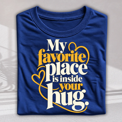 A folded royal blue t-shirt with a graphic print that reads 'My favorite place is inside your hug.' in white and yellow lettering with heart outlines, displayed on a light gray textured background.