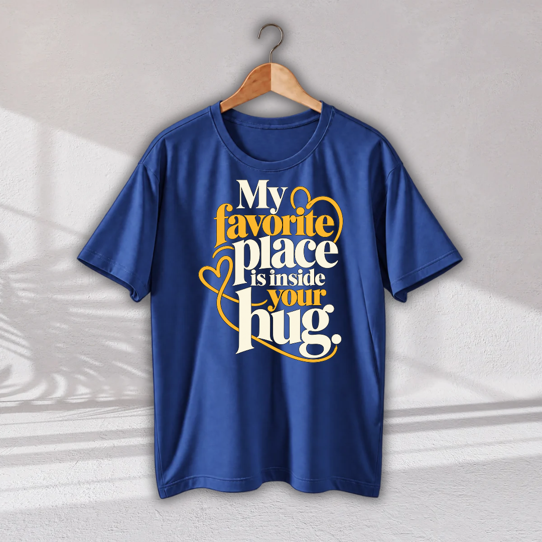 A royal blue short-sleeve t-shirt hanging on a wooden hanger against a light gray wall. The shirt features a graphic print on the front with the text 'My favorite place is inside your hug.