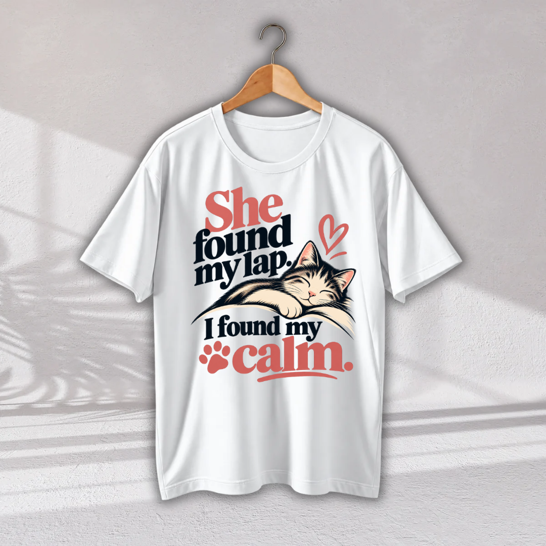 A white short-sleeve t-shirt hanging on a wooden hanger against a light gray wall. The shirt features a graphic print of a sleeping cat on a person's lap with the text 'She found my lap.