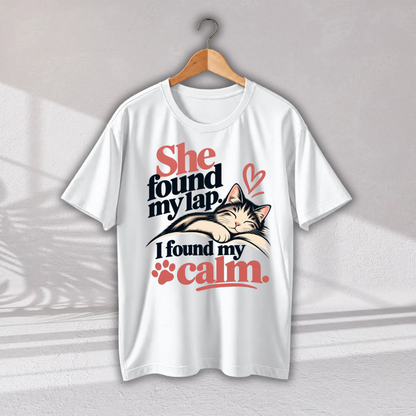 A white short-sleeve t-shirt hanging on a wooden hanger against a light gray wall. The shirt features a graphic print of a sleeping cat on a person's lap with the text 'She found my lap.