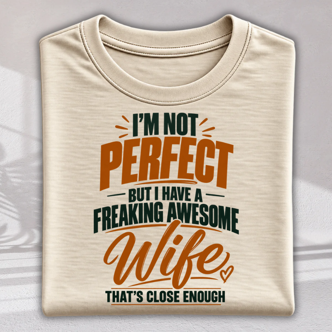 A folded cream-colored t-shirt with a humorous graphic print that reads 'I'M NOT PERFECT BUT I HAVE A FREAKING AWESOME Wife THAT'S CLOSE ENOUGH' in orange and dark green lettering, set against a light gray background.
