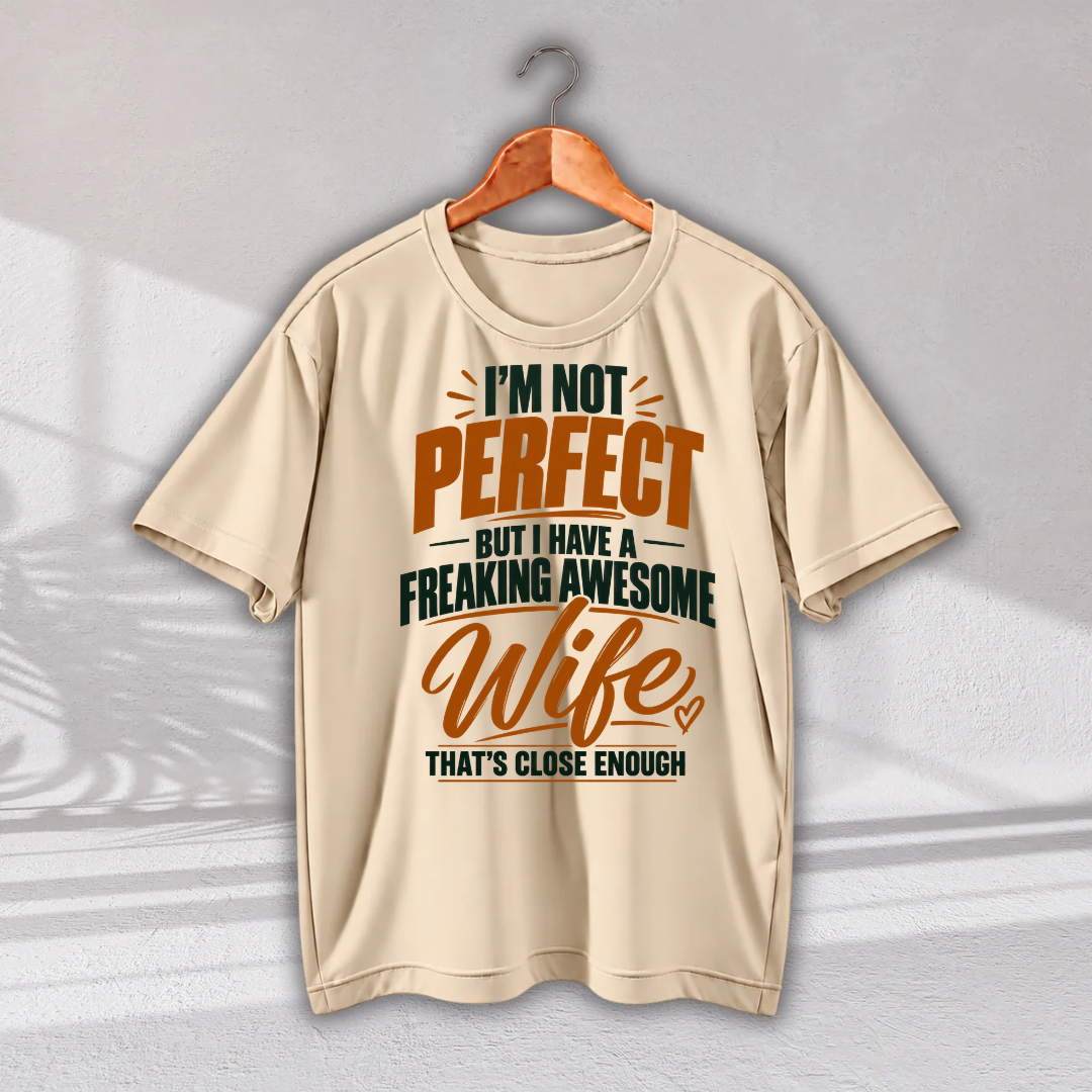 A beige short-sleeve t-shirt hanging on a wooden hanger against a light gray wall. The shirt features a humorous graphic print in orange and dark green text that reads: 'I'M NOT PERFECT BUT I HAVE A FREAKING AWESOME Wife THAT'S CLOSE ENOUGH'.