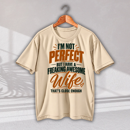 A beige short-sleeve t-shirt hanging on a wooden hanger against a light gray wall. The shirt features a humorous graphic print in orange and dark green text that reads: 'I'M NOT PERFECT BUT I HAVE A FREAKING AWESOME Wife THAT'S CLOSE ENOUGH'.