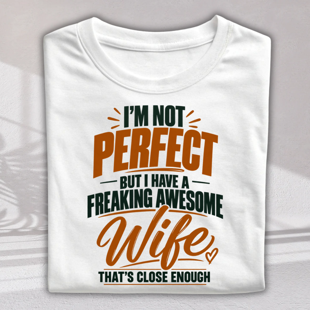 A folded white short-sleeve T-shirt with a humorous printed slogan on the front: 'I'M NOT PERFECT BUT I HAVE A FREAKING AWESOME Wife THAT'S CLOSE ENOUGH' in orange and dark green lettering, shown against a light gray background.