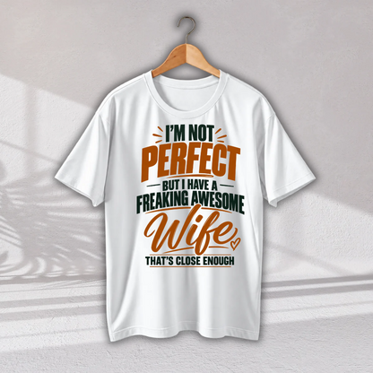 A white short-sleeve t-shirt hanging on a wooden hanger against a light gray wall. The shirt features a graphic text print that reads 'I'M NOT PERFECT BUT I HAVE A FREAKING AWESOME Wife THAT'S CLOSE ENOUGH' in orange, dark green, and black lettering.
