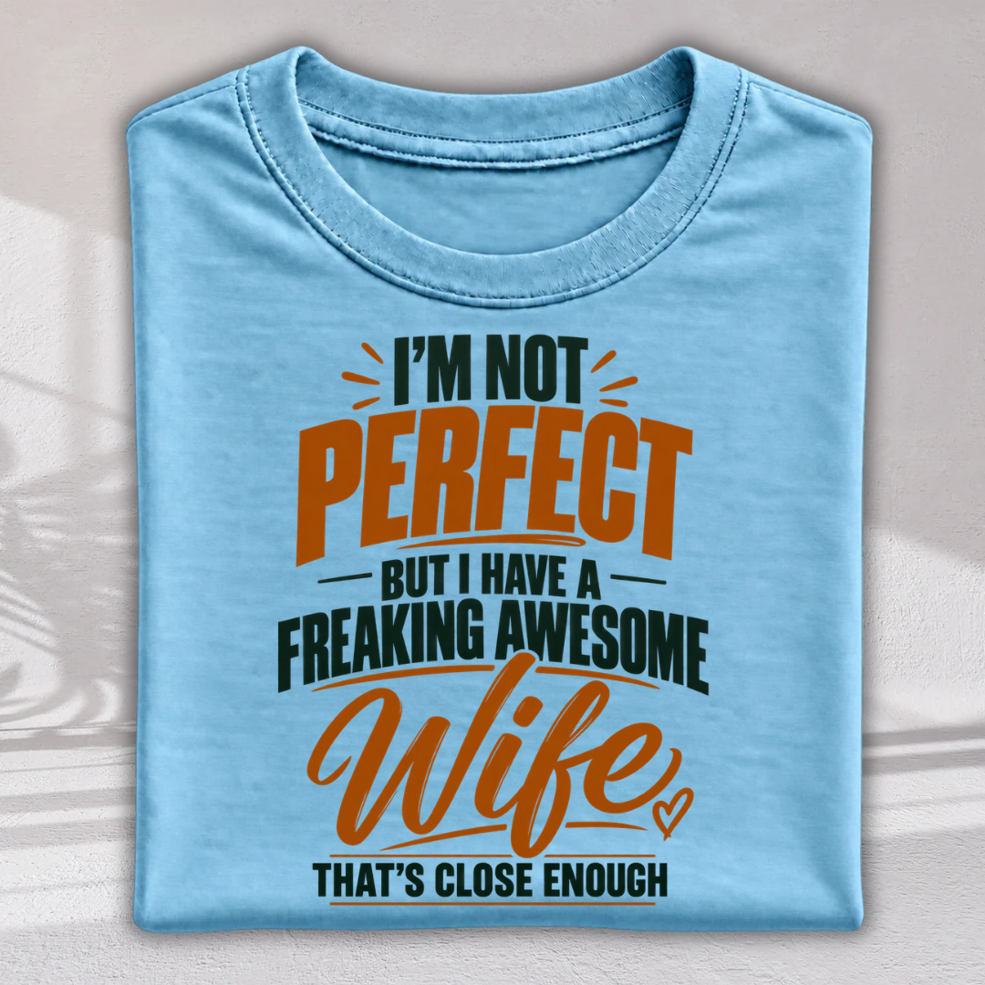 A folded light blue t-shirt with a humorous graphic print that reads 'I'M NOT PERFECT BUT I HAVE A FREAKING AWESOME Wife THAT'S CLOSE ENOUGH' in orange and dark green lettering, displayed against a neutral background with soft shadow overlays.