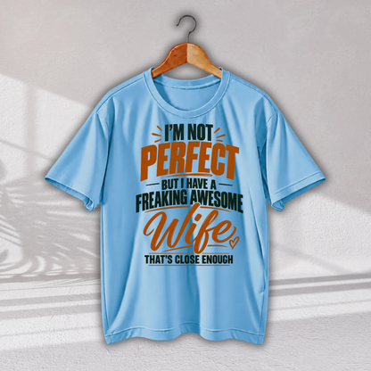 A light blue short-sleeve t-shirt hanging on a wooden hanger against a light gray wall. The shirt features a graphic text print that reads 'I'M NOT PERFECT BUT I HAVE A FREAKING AWESOME Wife THAT'S CLOSE ENOUGH' in orange and dark teal lettering.