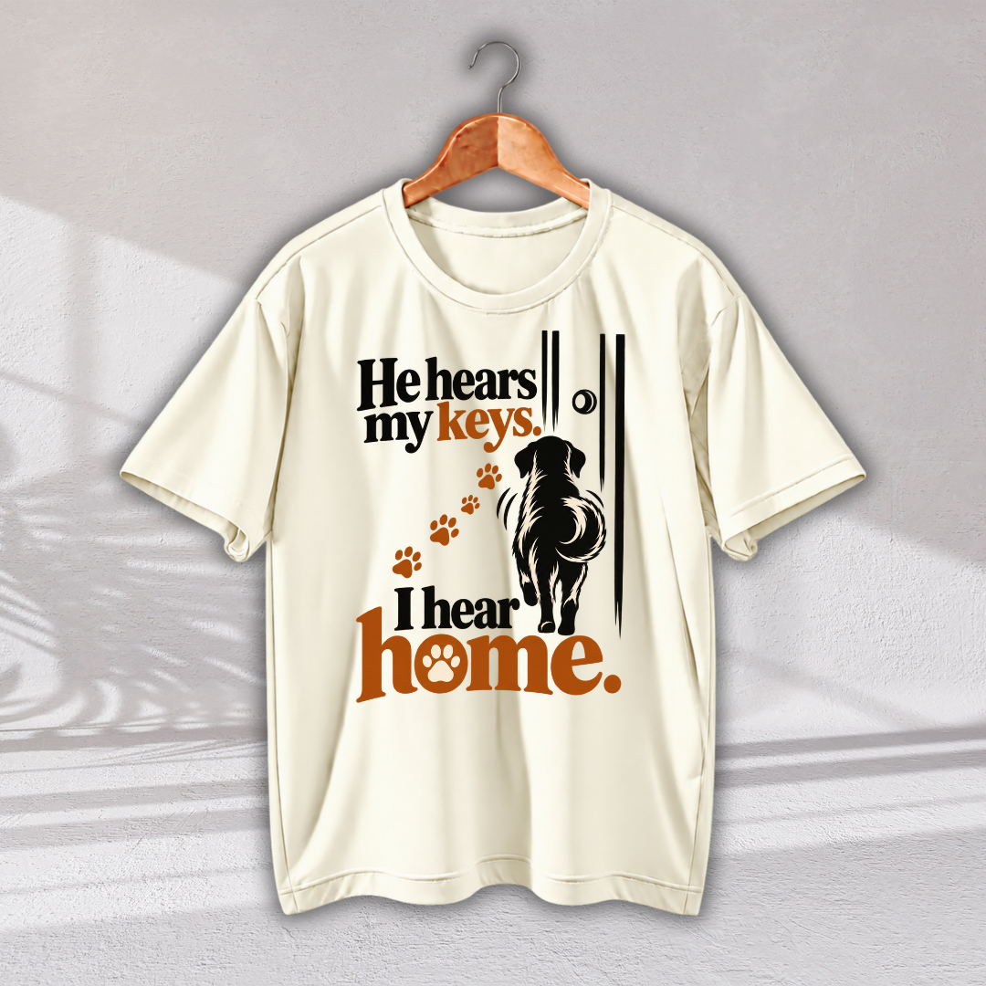 An off-white short-sleeve t-shirt hanging on a wooden hanger against a light gray wall. The shirt features a graphic print with the text 'He hears my keys I hear home.