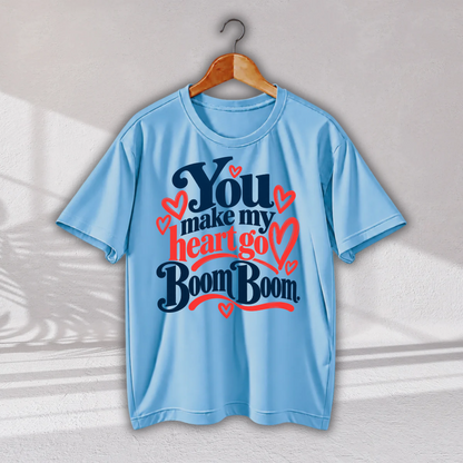 A light blue short-sleeve t-shirt hanging on a wooden hanger against a light gray wall. The shirt features a graphic print on the front with the text 'You make my heart go Boom Boom' in dark blue and red lettering, accented with small red hearts.
