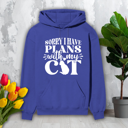 Blue hoodie with 'Sorry I have plans with my cat' text and cat silhouette, placed on a wooden floor with flowers and a plant.
