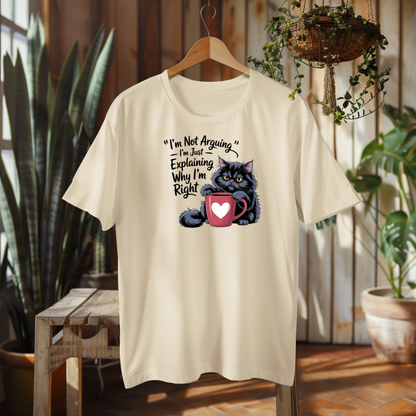 Beige t-shirt with a graphic of a cat holding a mug and text, hanging on a wooden hanger in a room with plants.
