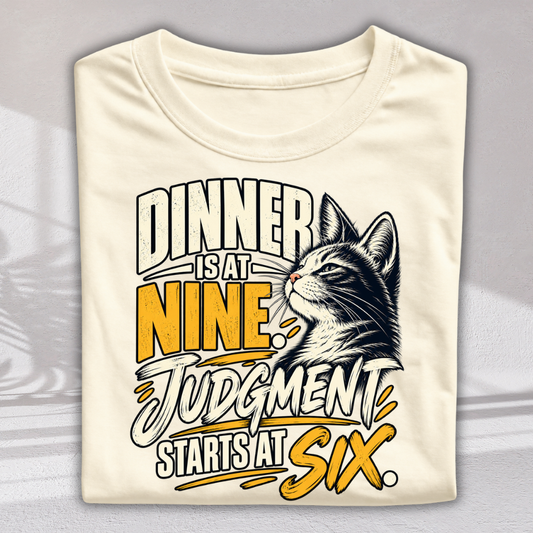 A folded cream-colored t-shirt with a graphic print featuring a cat and the text 'DINNER IS AT NINE. JUDGMENT STARTS AT SIX.