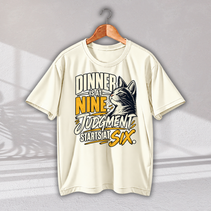 An off-white short-sleeve t-shirt on a wooden hanger, featuring a graphic print of a cat's head and the text 'DINNER IS AT NINE, JUDGMENT STARTS AT SIX.' in yellow, black, and white.