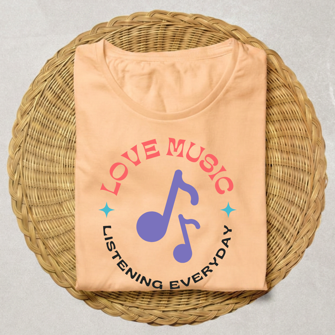 A peach-colored t-shirt with a graphic print reading 'LOVE MUSIC' in pink and 'LISTENING EVERYDAY' in black, featuring a purple musical note in the center, displayed on a round woven wicker tray.