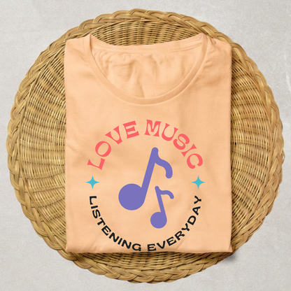 A peach-colored t-shirt with a graphic print reading 'LOVE MUSIC' in pink and 'LISTENING EVERYDAY' in black, featuring a purple musical note in the center, displayed on a round woven wicker tray.