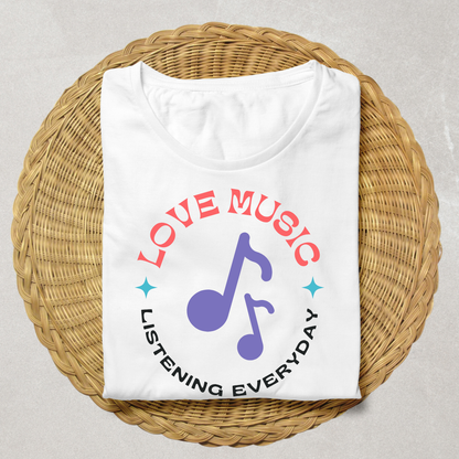 A folded white t-shirt with a colorful graphic print reading 'LOVE MUSIC LISTENING EVERYDAY' around a purple music note, displayed on a round woven straw mat against a light gray background.