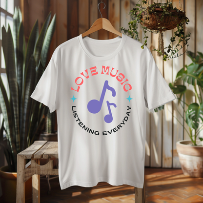 A white short-sleeve t-shirt hanging on a wooden hanger in a rustic indoor setting. The shirt features a graphic print with the words 'LOVE MUSIC' in coral pink and 'LISTENING EVERYDAY' in black, surrounding a purple musical note icon.