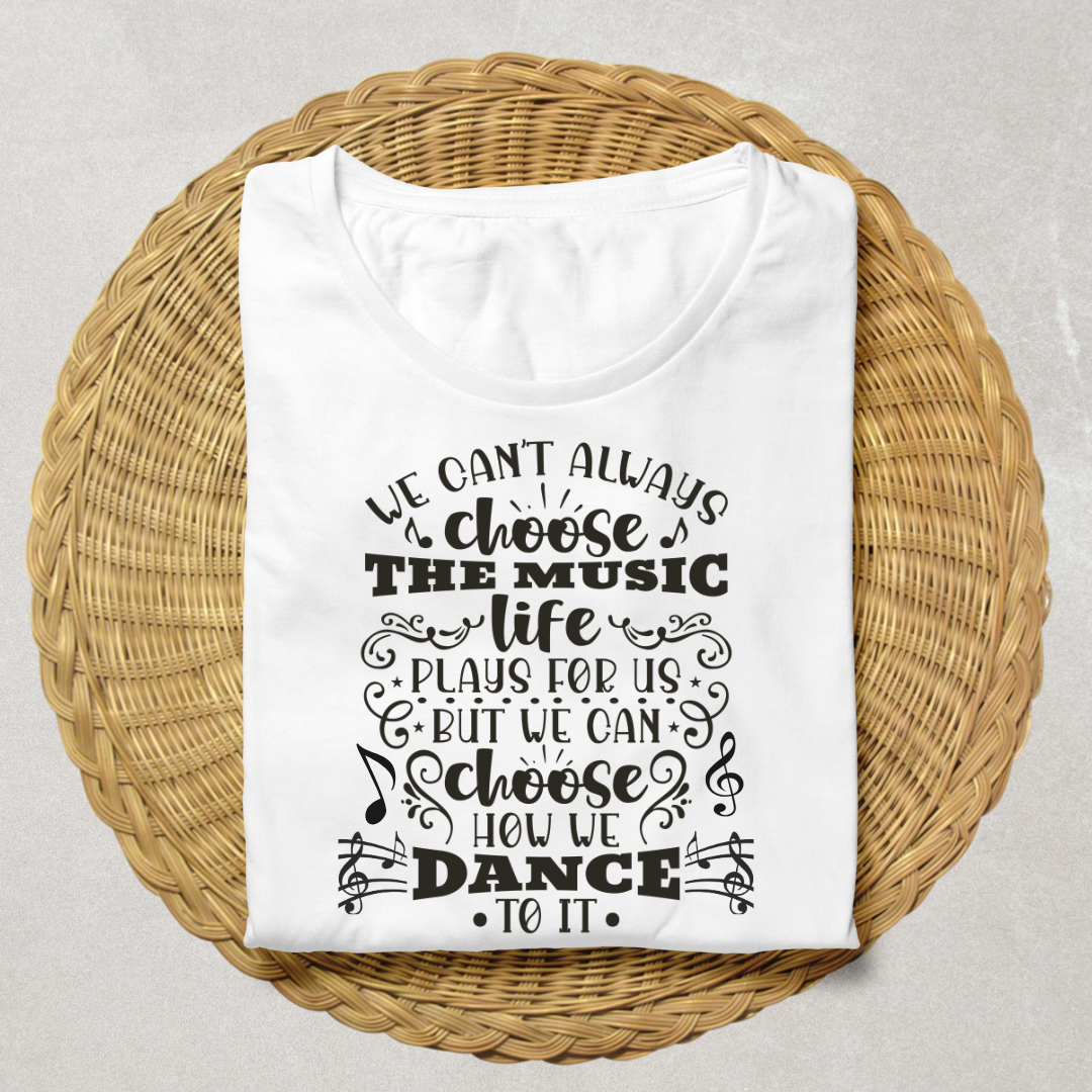 A white short-sleeve T-shirt with black printed text "We can't always choose the music life plays for us but we can choose how we dance to it" and musical note graphics, laid flat on a round woven wicker tray against a light neutral background.
