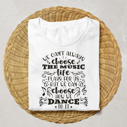 A white short-sleeve T-shirt with black printed text "We can't always choose the music life plays for us but we can choose how we dance to it" and musical note graphics, laid flat on a round woven wicker tray against a light neutral background.